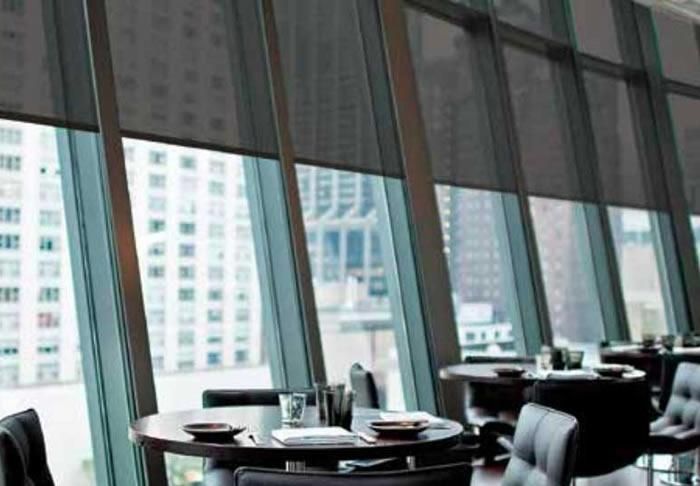 Lutron cable-guided shades installed on large, slanted windows in a modern dining area with cityscape view in the background.