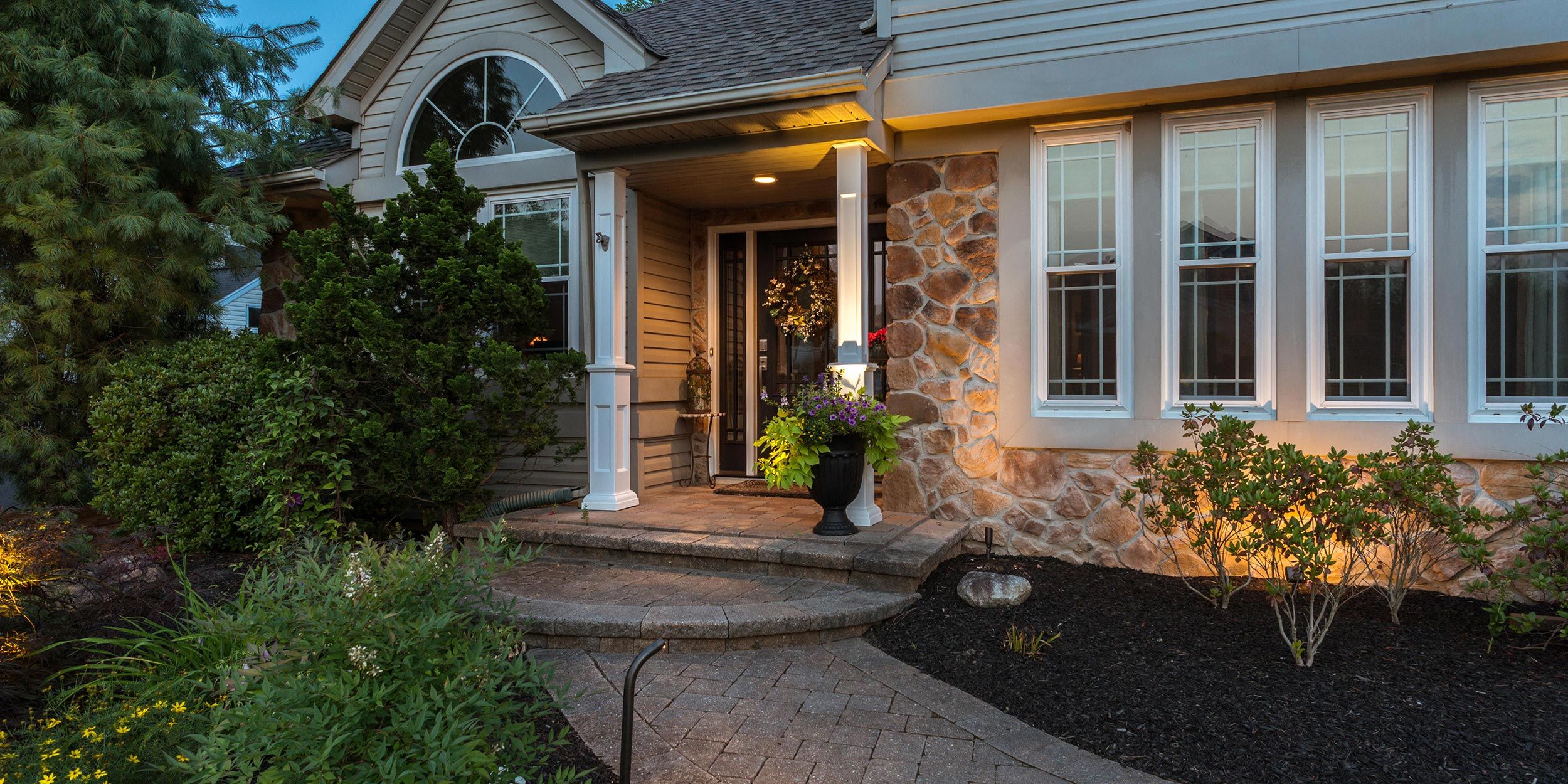 A welcoming front porch with a mix of stone and wood textures, warm lighting, and well-maintained shrubs flanking the entrance.